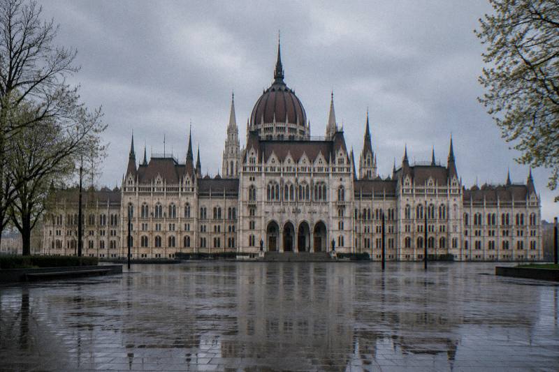 Mañana lluviosa en la amplia Plaza Kossuth de Budapest, Hungría, con la fachada terrestre del Parlamento, su cúpula central y las agujas neogóticas vistas de frente a través de una plaza ancha de pavimento oscurecido por la lluvia, algunos árboles sin hojas o a punto de brotar en los bordes y sin el río a la vista.