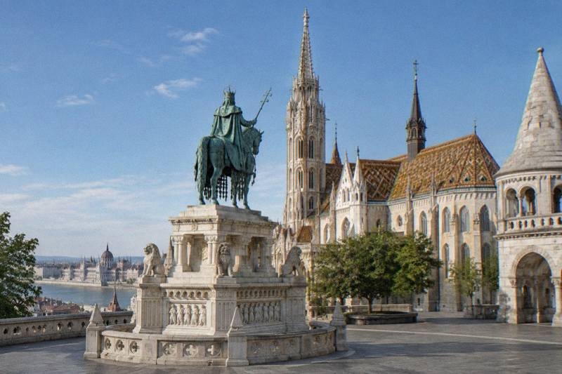 Crisp morning on the terrace of the Fisherman's Bastion in Budapest, Hungary, a bronze equestrian statue on a tall stone plinth with the tiled roof and spire of the Matthias Church rising behind it, empty paving, a blue sky.