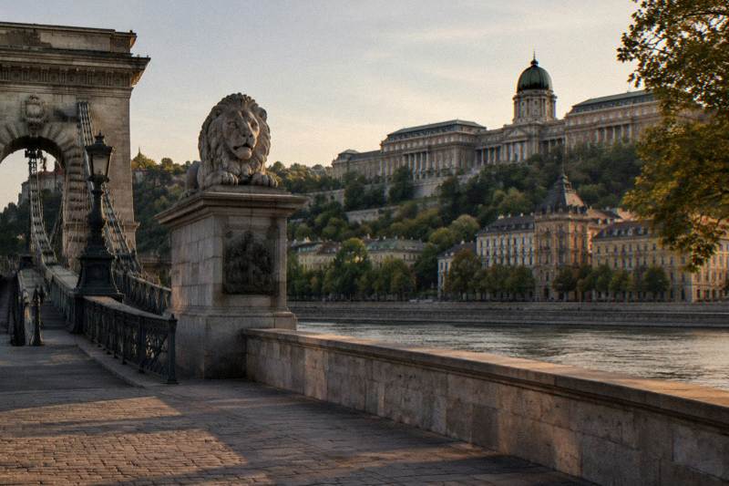 Tarde avanzada en el extremo de Buda del Puente de las Cadenas en Budapest, Hungría, con el macizo estribo de piedra y un león tallado en primer plano, mientras la arbolada ladera de la Colina del Castillo se alza tras él, largas sombras cálidas sobre el pavimento vacío.