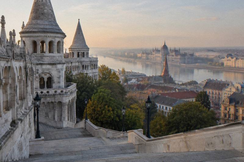 Early morning on the Fisherman's Bastion in Budapest, Hungary, the white limestone terraces, arcades and conical turrets stepping down a staircase, soft haze over the Danube far below, empty stone steps in the foreground.