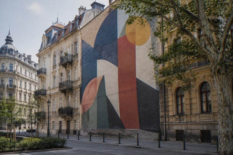 Late-morning light on a tall brick firewall in the Jewish Quarter of Budapest, Hungary, covered by a large abstract painted mural in bold geometric shapes and no lettering, plain apartment facades and a plane tree beside it, empty street.
