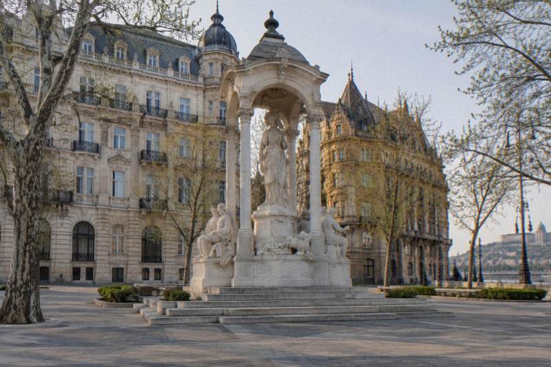 Spring morning on Vorosmarty Square in Budapest, Hungary, a large white marble monument group beneath a stone canopy in the middle of the frame, tall cream Eclectic facades and bare-to-budding plane trees around it, empty paving.