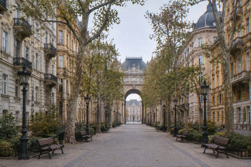 Soft overcast morning on the Madach Imre promenade in Budapest, Hungary, a long straight avenue of plane trees between tall cream-and-ochre Eclectic apartment blocks, a monumental arched gateway closing the view, empty benches.