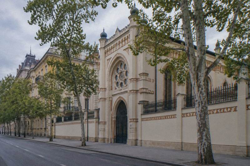 Overcast afternoon at the Kazinczy Street Orthodox Synagogue in Budapest, Hungary, a pale Art Nouveau facade with a tall arched entrance and decorative brick bands, a high plain wall and plane trees along the empty street.