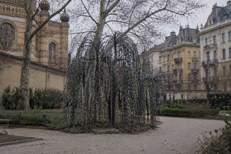 Grey afternoon in the garden behind the Great Synagogue in Budapest, Hungary, a large weeping-willow sculpture of dark metal leaves in the middle of the frame with a pale brick wall and bare branches behind it, gravel path in the foreground.