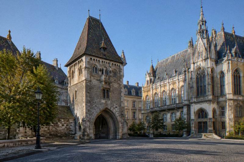 Mañana despejada en la Puerta de Viena, en el Distrito del Castillo de Budapest, Hungría, con la torre de piedra de la puerta y su tejado empinado de tejas, junto a un edificio neogótico alto, plaza empedrada en primer plano y cielo azul.