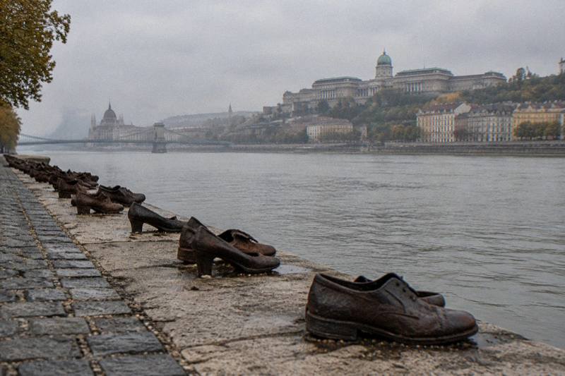 Grey afternoon on the Danube embankment in Budapest, Hungary, a long row of rusted cast-iron shoes along the stone edge of the quay, the grey-green river beyond and the far Buda bank blurred in mist.