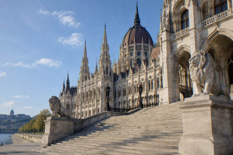 Mañana soleada a los pies del Parlamento en Budapest, Hungría: una amplia escalinata de piedra flanqueada por leones de piedra que lleva hasta la ornamentada fachada neogótica, de piedra clara y altas aguja contra un cielo azul