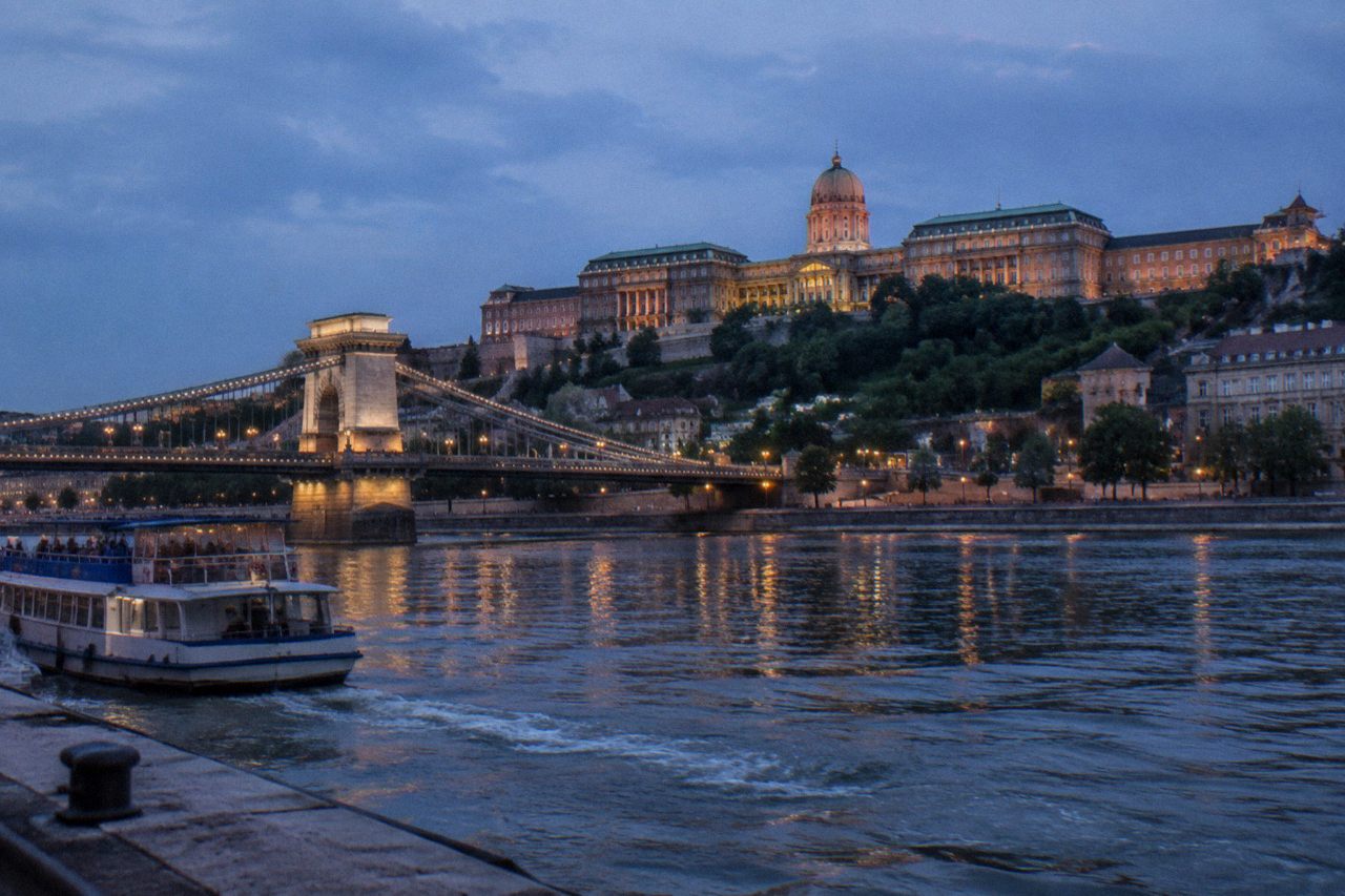 Buda Castle on Castle Hill above the Danube at dusk, with the Chain Bridge and Pest skyline in the background
